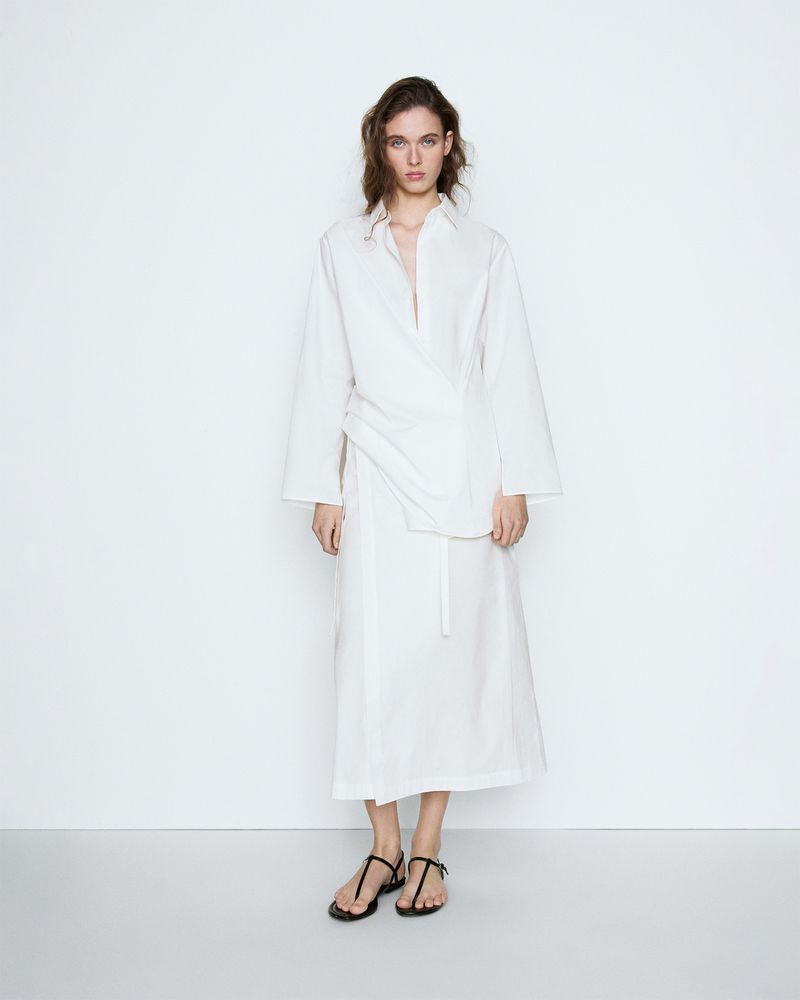Model wearing a long white wrap shirt dress and minimalist black strap sandals against a clean white studio backdrop.