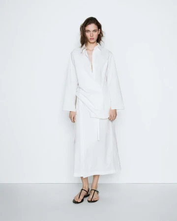 Model wearing a long white wrap shirt dress and minimalist black strap sandals against a clean white studio backdrop.