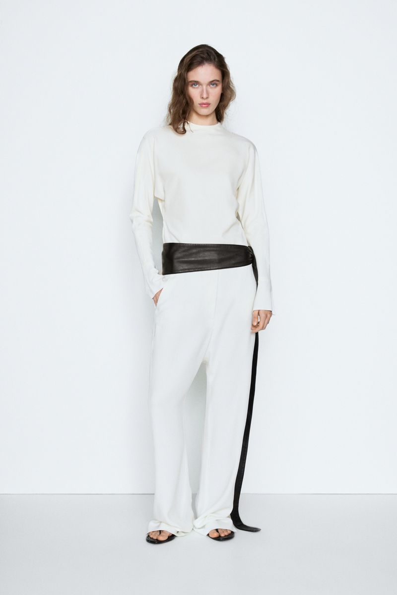 Model standing against white backdrop wearing white long-sleeve top and white trousers with a wide dark leather belt.