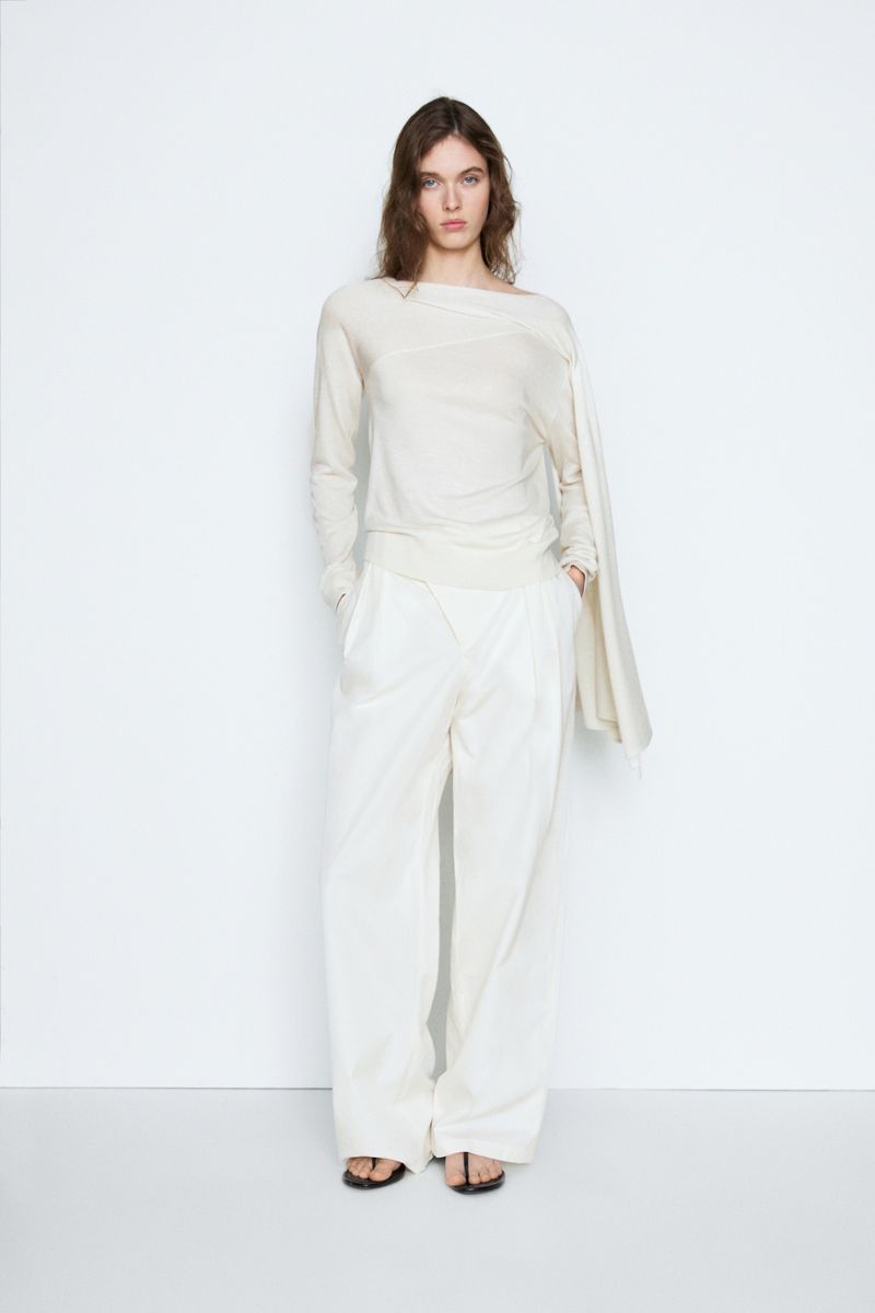 Model posing in an off-white asymmetrical long-sleeve top and matching wide-leg trousers against a white backdrop.