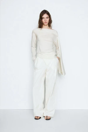 Model posing in an off-white asymmetrical long-sleeve top and matching wide-leg trousers against a white backdrop.