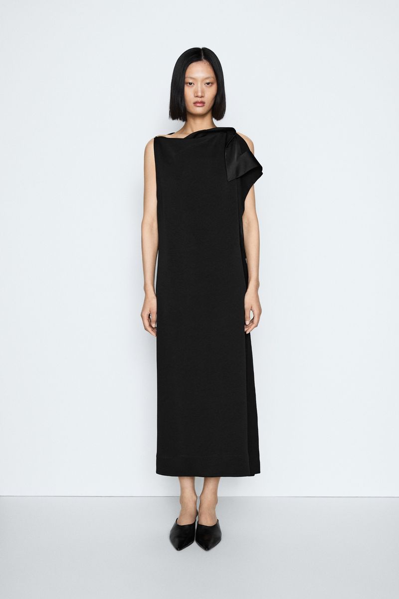 Model with dark bob haircut wearing a long black dress standing against a light grey seamless studio background.