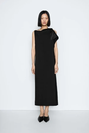 Model with dark bob haircut wearing a long black dress standing against a light grey seamless studio background.
