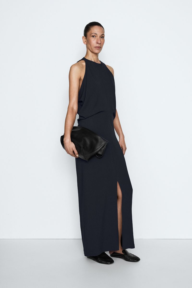 Model Liset Mitjan posing in a sleeveless dark navy maxi dress with a front slit and black leather clutch against a white studio sweep.