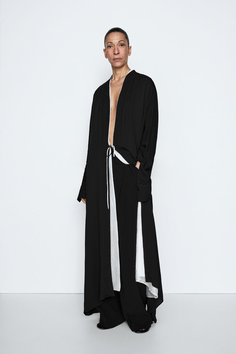 Model Liset Mitjan posing in a flowing black robe coat with white lining and wide-leg trousers against a white seamless backdrop.