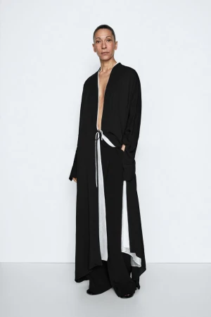 Model Liset Mitjan posing in a flowing black robe coat with white lining and wide-leg trousers against a white seamless backdrop.