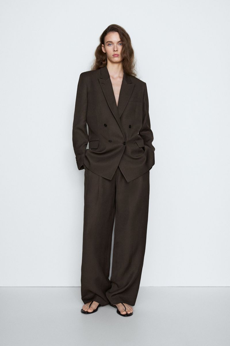 Model posing in an oversized dark brown tailored pantsuit with black flat thong sandals against a white studio backdrop.