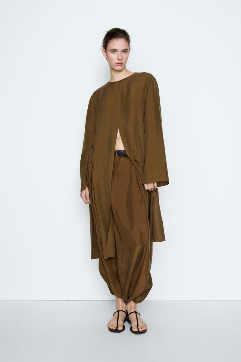 Model Liset Mitjan posing in an olive brown draped long tunic set and black flat sandals against a white studio background.