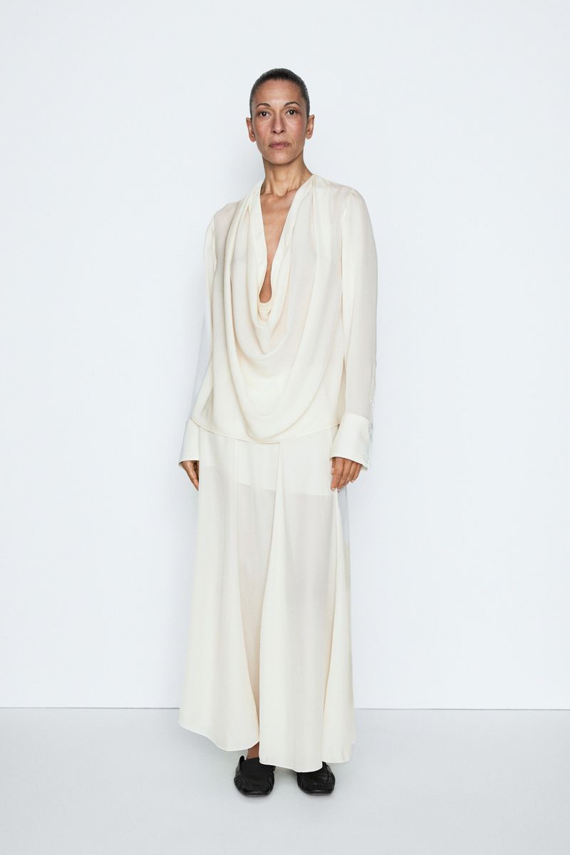 Model posing in an off-white draped cowl-neck maxi dress with black shoes against a white backdrop.