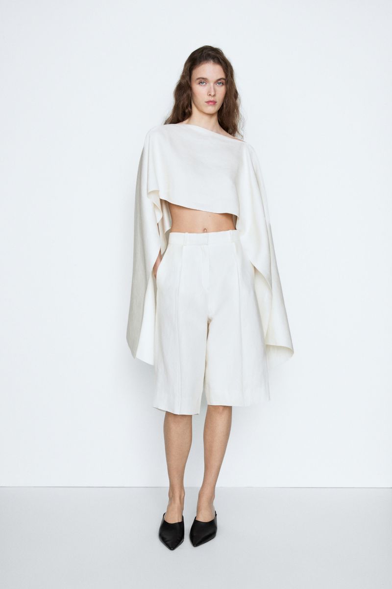 Model posing in an off-white structured cape crop top and matching tailored shorts with black mules against a white studio sweep.