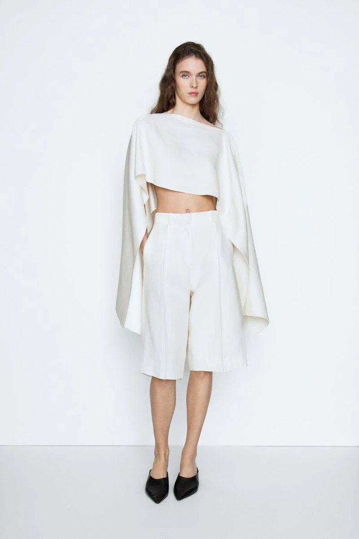 Model posing in an off-white structured cape crop top and matching tailored shorts with black mules against a white studio sweep.