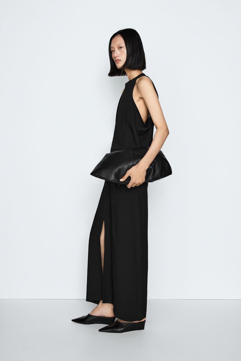 Model Yilan Hua standing in profile wearing a black sleeveless maxi dress and holding an oversized black clutch against a white backdrop.