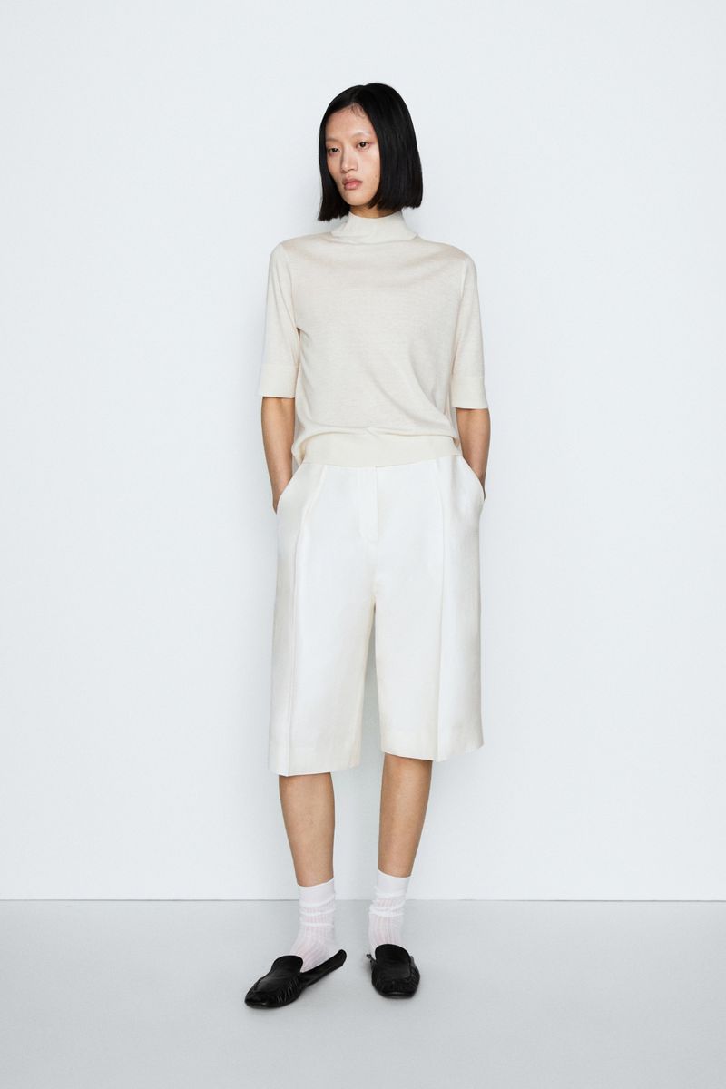 Model Yilan Hua posing in a cream mock neck top and tailored off-white bermuda shorts against a white backdrop.