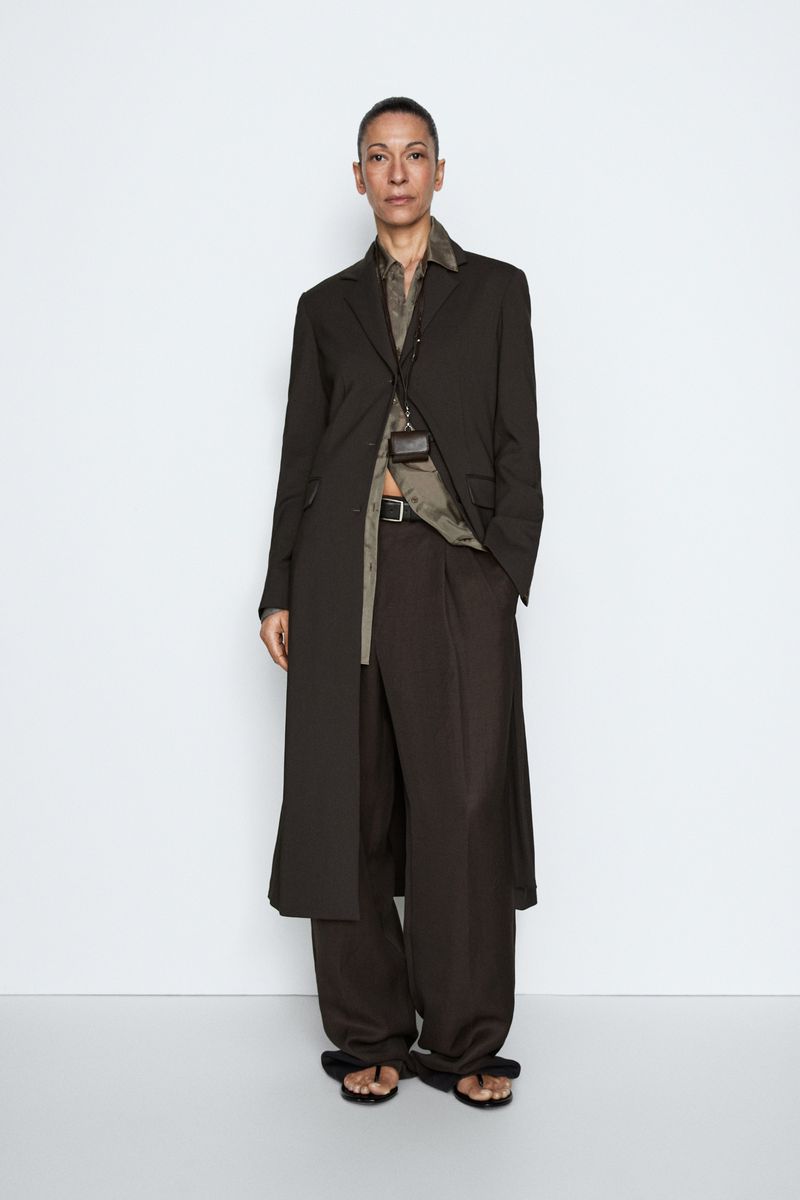 Model wearing a long dark brown tailored coat and matching wide-leg trousers against a white seamless studio background.