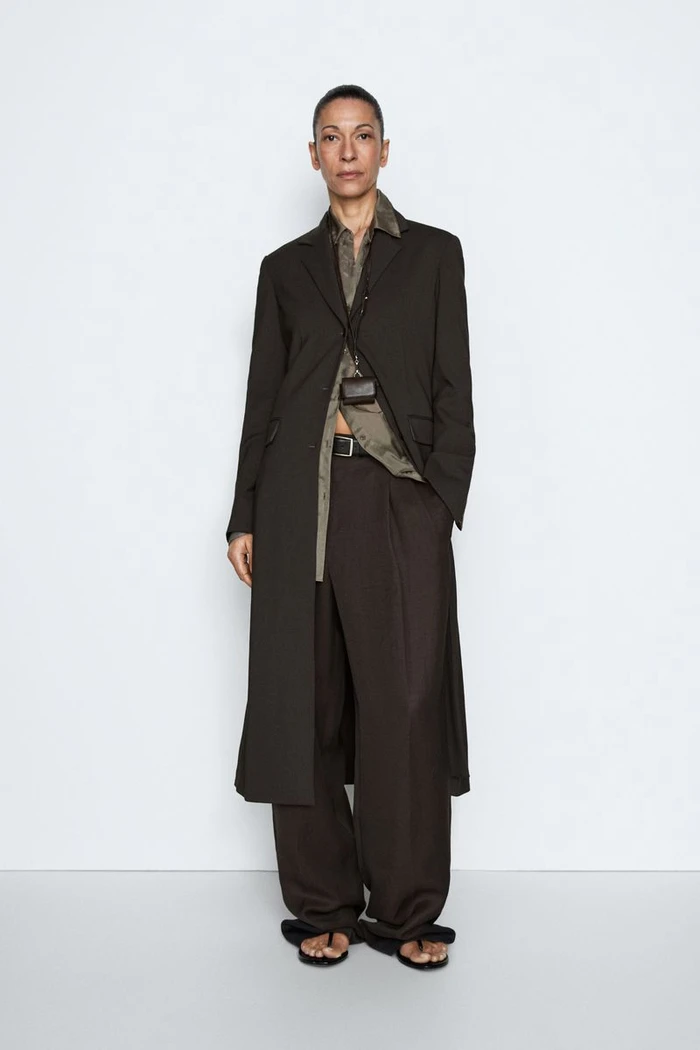 Model wearing a long dark brown tailored coat and matching wide-leg trousers against a white seamless studio background.