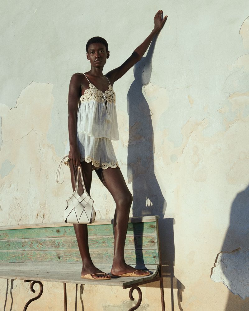Model Fatou Kebbeh wearing a white lace camisole and matching shorts standing on a bench against a rustic wall.
