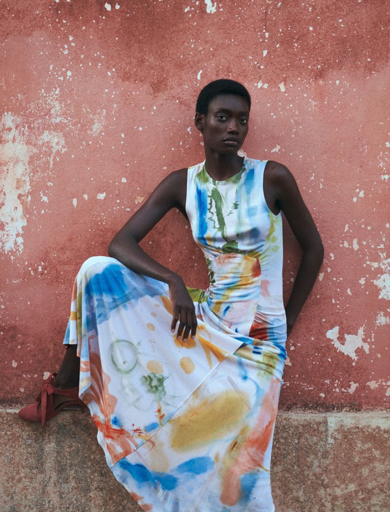 Model posing in a colorful abstract watercolor sleeveless dress against a distressed terracotta textured wall.