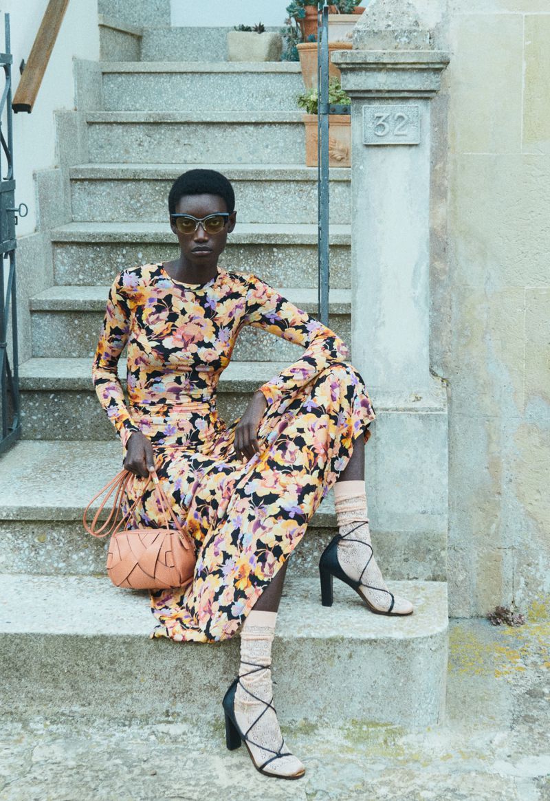 Model wearing a floral print dress and black sunglasses sitting on stone outdoor steps holding a small tan bag