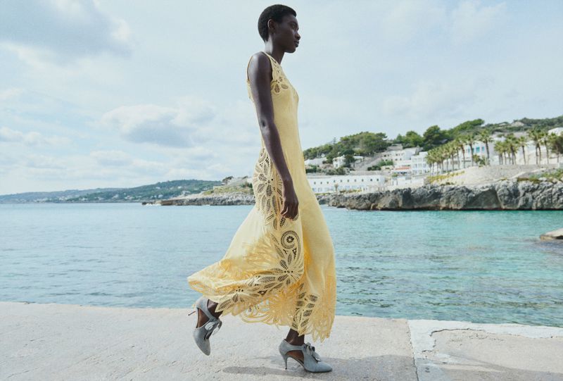 Model Fatou Kebbeh walks along a seaside pier in an intricate yellow cutout lace maxi dress for Ulla Johnson.