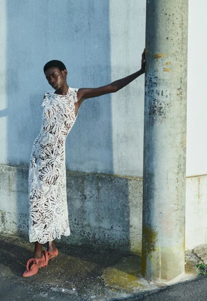 Model Fatou Kebbeh poses in a sleeveless white lace dress and terracotta heels leaning against a concrete post outdoors.