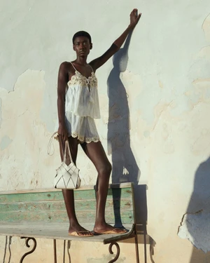 Model Fatou Kebbeh wearing a white lace camisole and matching shorts standing on a bench against a rustic wall.