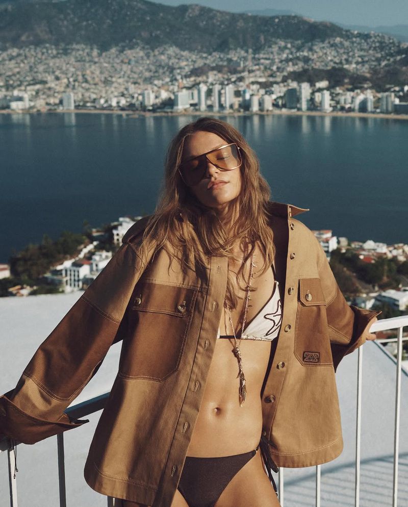 Model Taylor D wears an open tan utility jacket, sunglasses, and a bikini overlooking a coastal city from a balcony.