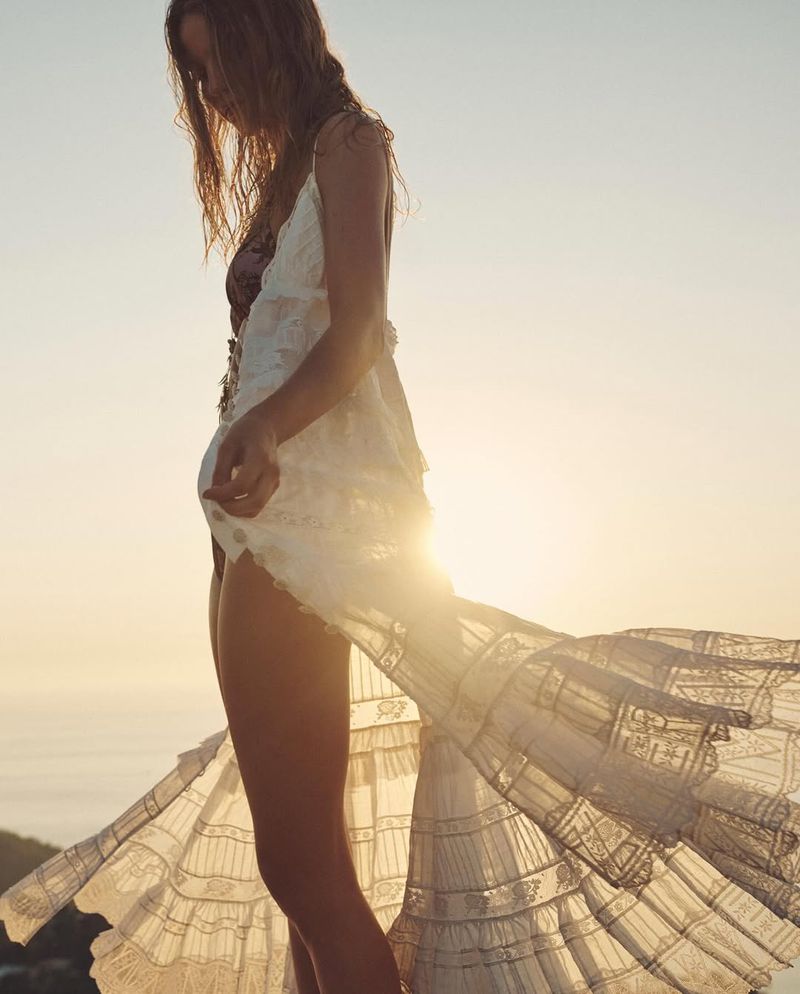 Model wearing a sheer, flowing white lace dress backlit by the golden sun against an open horizon.