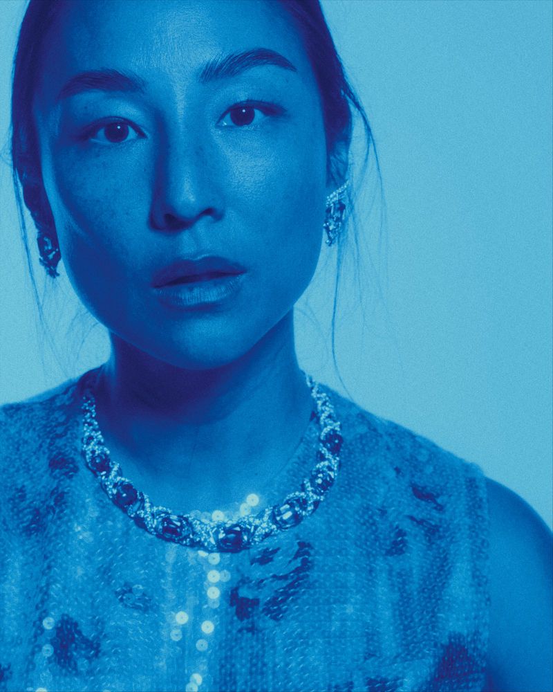 Close-up portrait of Greta Lee illuminated under cool blue light wearing a beaded statement necklace and textured top.