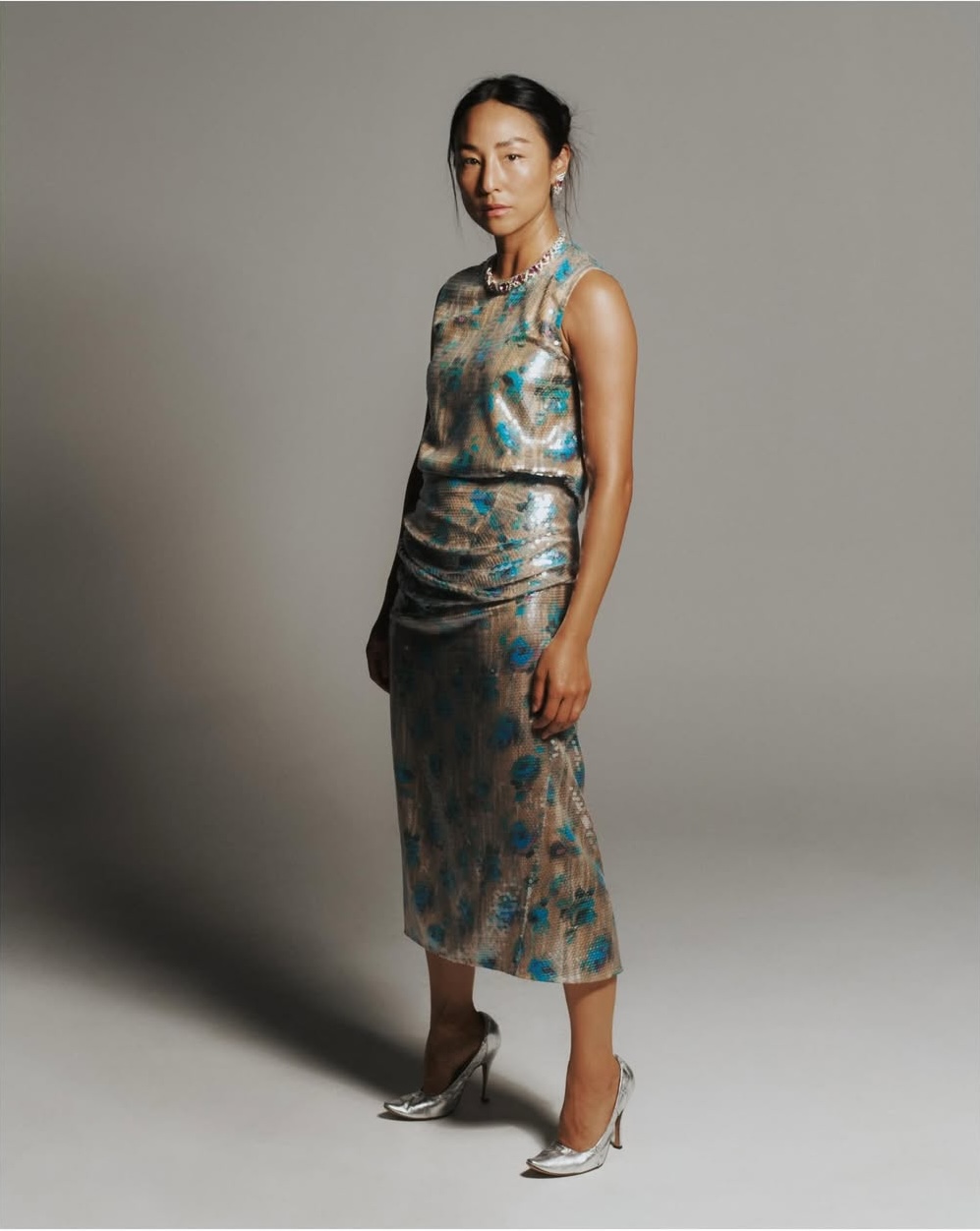 Greta Lee standing in a printed metallic sleeveless midi dress and silver heels against a grey studio backdrop.