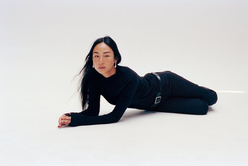 Actor Greta Lee wearing a black long-sleeve top and black trousers lying on a white seamless studio floor.