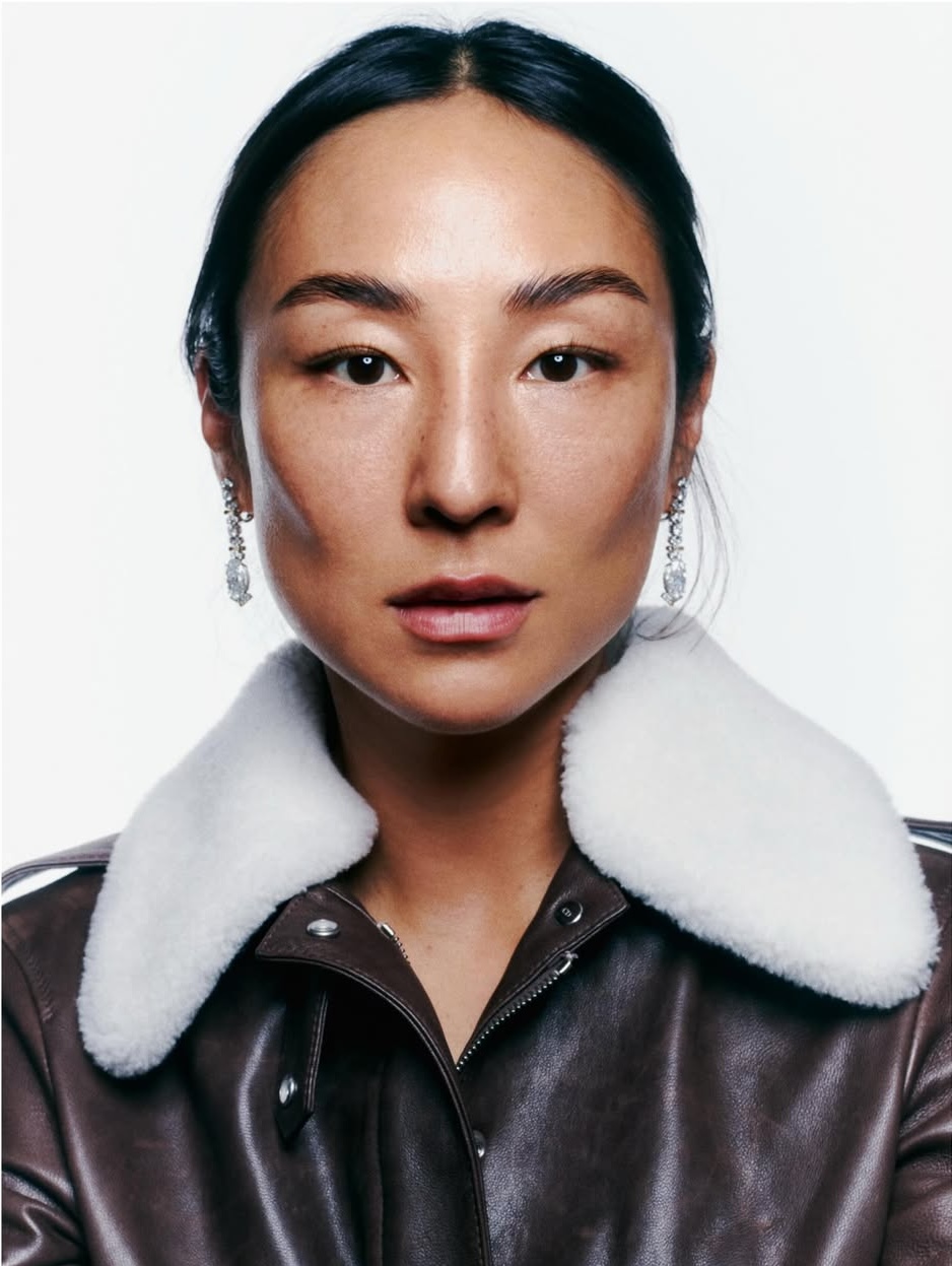 Close-up portrait of actor Greta Lee wearing a brown leather jacket with a white shearling collar and drop earrings against a white background.