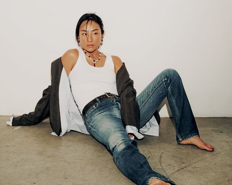 Greta Lee seated on a concrete floor wearing a white tank top and blue jeans, with an oversized jacket draped off her shoulders.