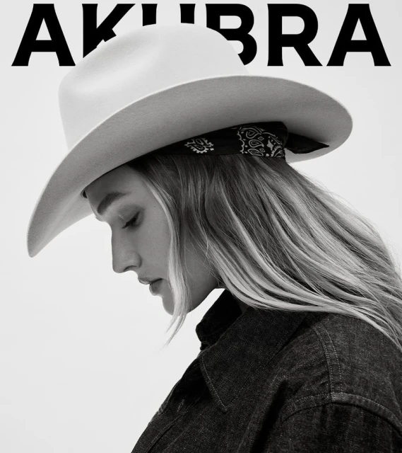 A woman wearing a white cowboy hat and denim jacket poses in a studio setting with soft lighting, showcasing fashion and beauty.