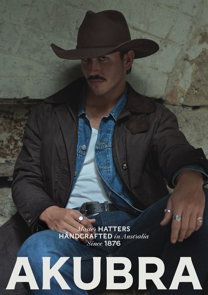 Man with mustache wearing brown hat and jacket, seated indoors against a stone wall, promoting Akubra hats in a medium shot.
