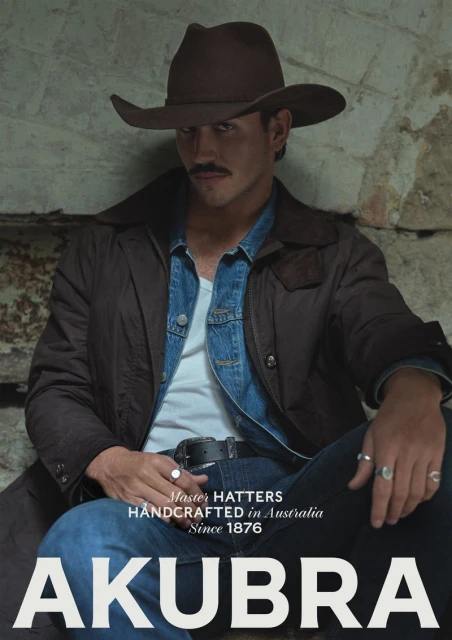 Man with mustache wearing brown hat and jacket, seated indoors against a stone wall, promoting Akubra hats in a medium shot.