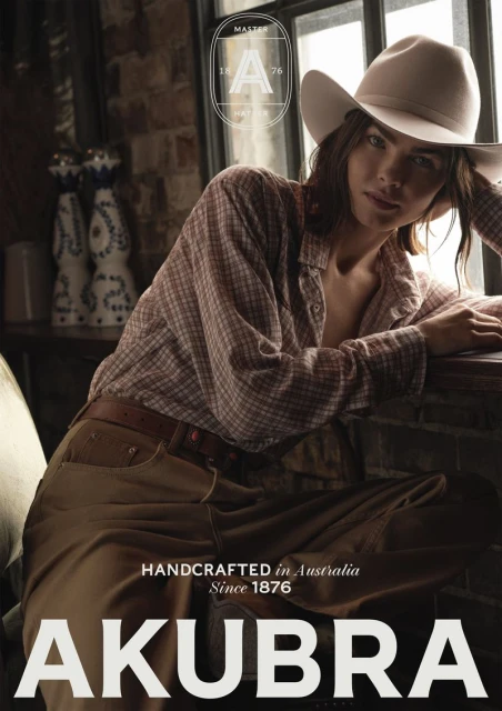 Woman wearing a cowboy hat and a plaid shirt leans against a windowsill with bottles on a shelf. The background is a brick wall.