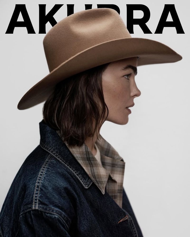 A woman poses in profile, wearing a sand-colored Akubra hat and a blue denim jacket with a plaid collar, against a neutral background.