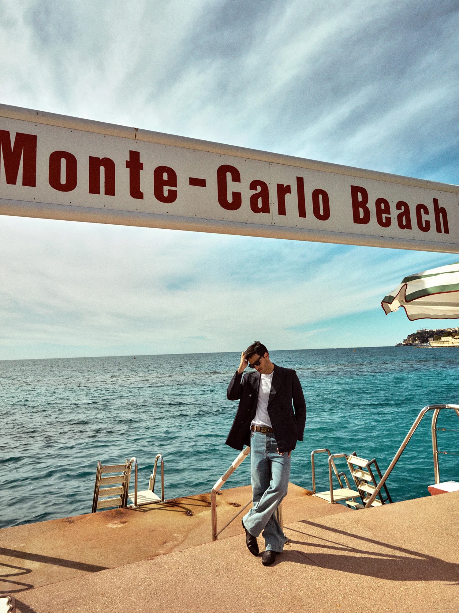 Carlos Sainz in a blazer and jeans posing beneath a Monte-Carlo Beach sign with the sea in the background.