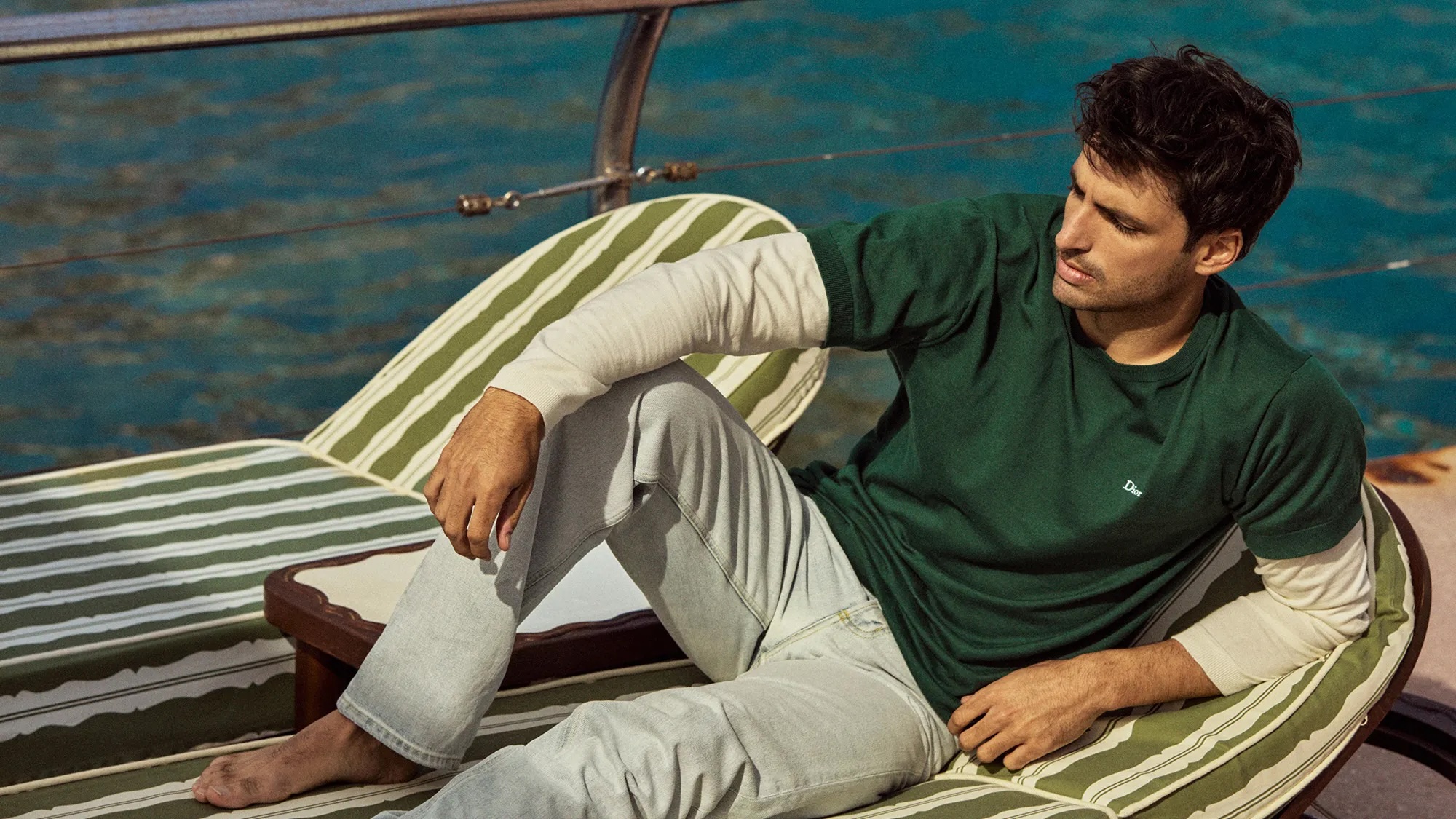 Carlos Sainz reclined on a striped sun lounger aboard a boat wearing a green t-shirt over long sleeves and light wash jeans.
