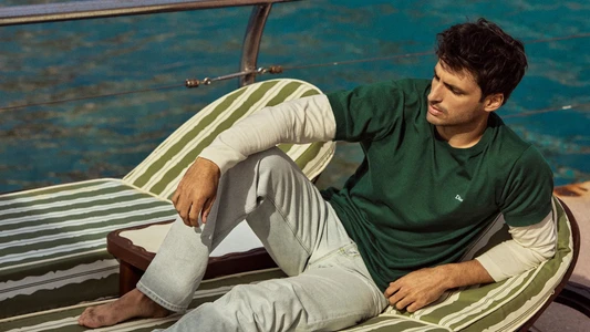 Carlos Sainz reclined on a striped sun lounger aboard a boat wearing a green t-shirt over long sleeves and light wash jeans.