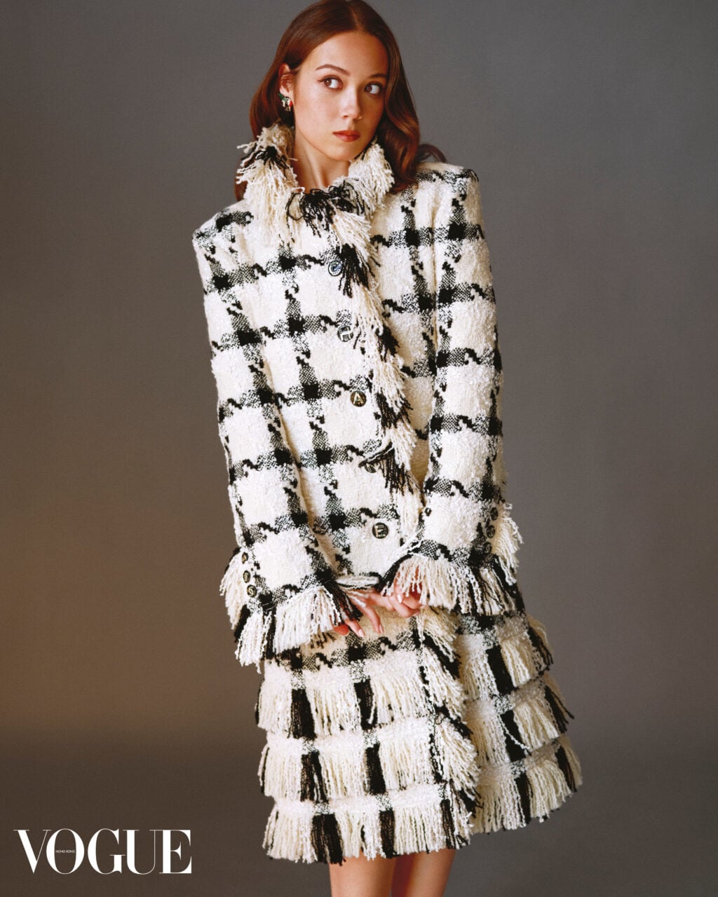 Laufey wearing a black and off-white plaid tweed coat with layered fringe trim against a neutral studio backdrop.