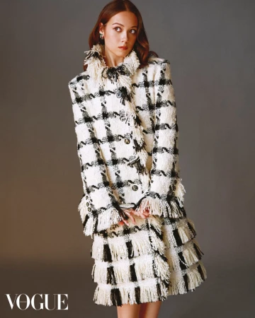 Laufey wearing a black and off-white plaid tweed coat with layered fringe trim against a neutral studio backdrop.