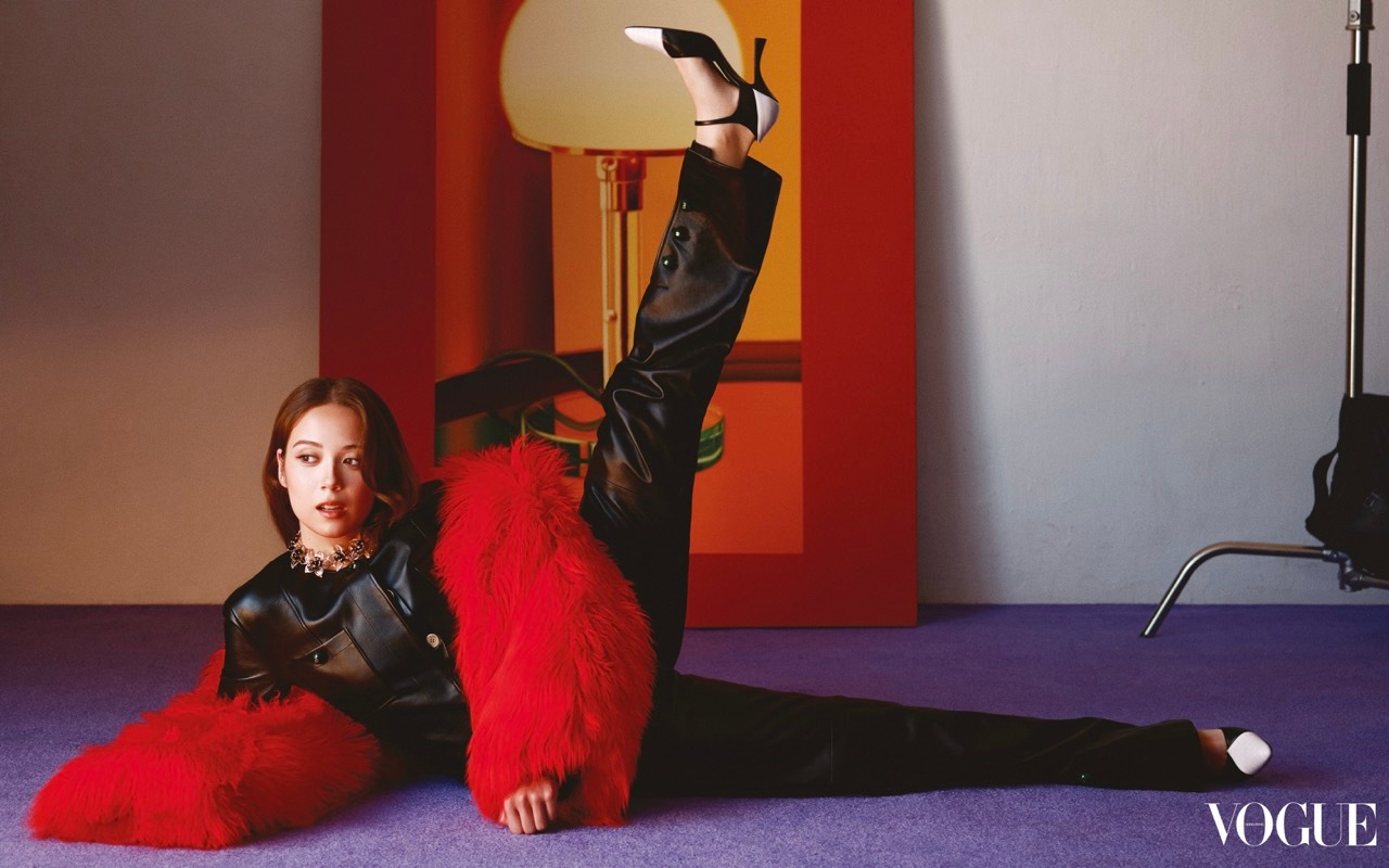 Musician Laufey lying on a purple floor with one leg raised, wearing a black leather outfit, bright red fur wrap, and two-tone heels.