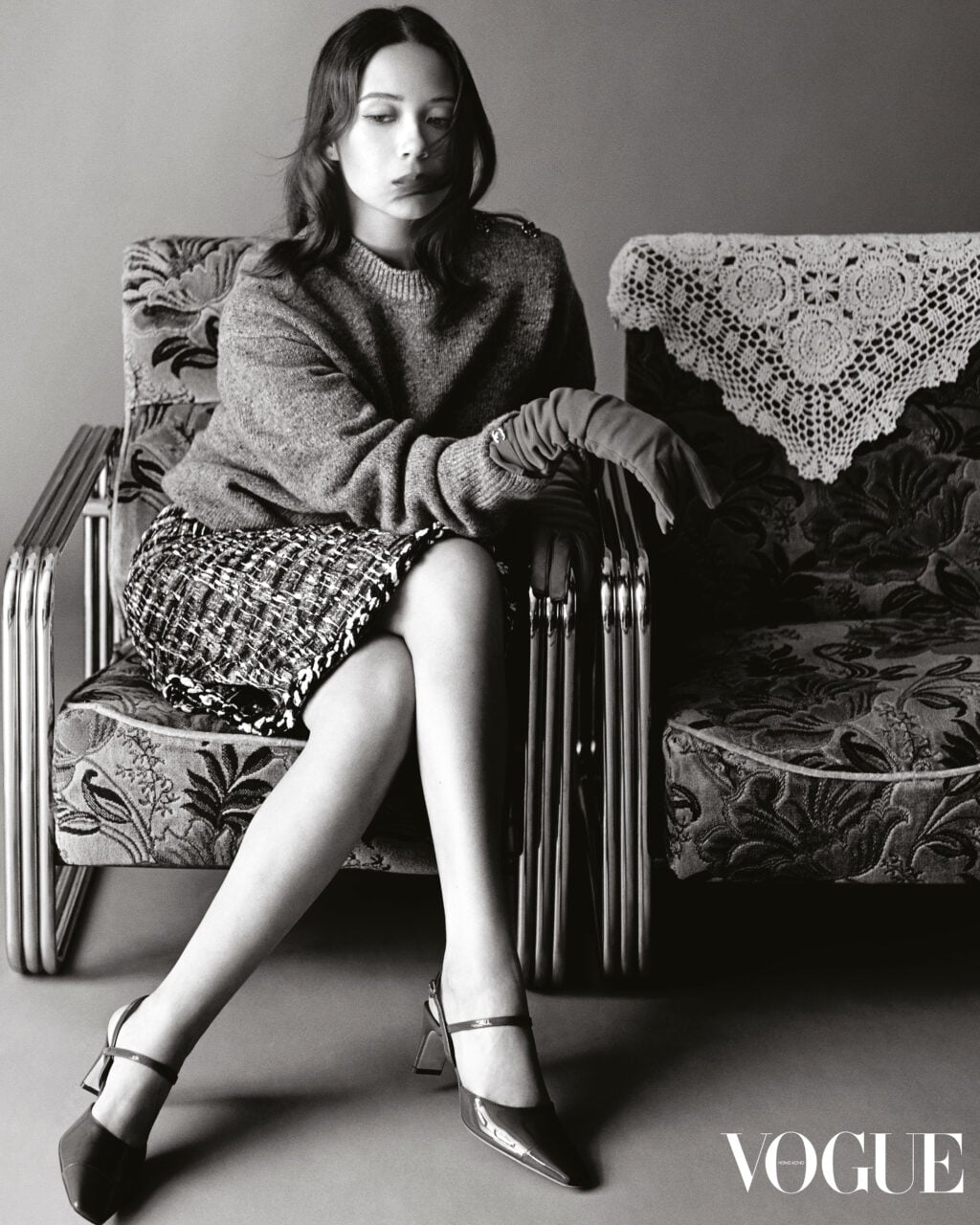 Laufey seated on an upholstered vintage armchair in a knit sweater, tweed skirt, and pointed slingback heels for Vogue Hong Kong.