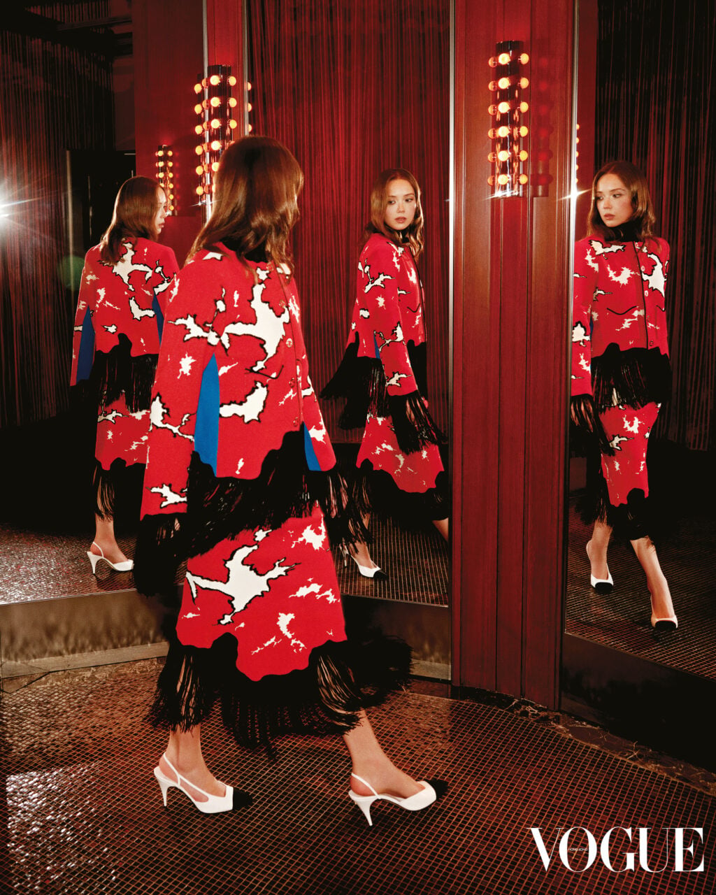 Laufey walks past mirrors wearing a red patterned coat dress with black fringe and white slingback heels in a red room.