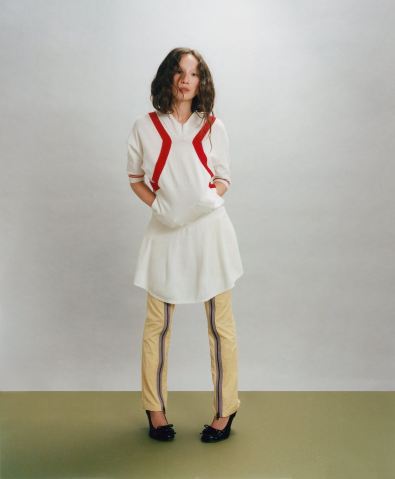 Havana Rose Liu posing in a white sporty tunic dress over pale yellow zippered trousers and black heels against a studio backdrop.