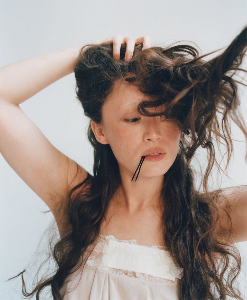 Havana Rose Liu holding long wavy dark hair with bobby pins in mouth, wearing a taped camisole against a white backdrop.