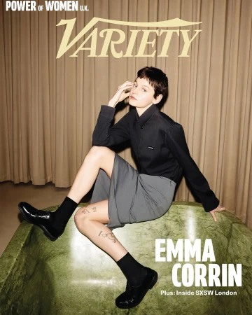 Portrait of actor Emma Corrin seated on a green bathtub wearing a dark shirt and grey skirt against draped curtains