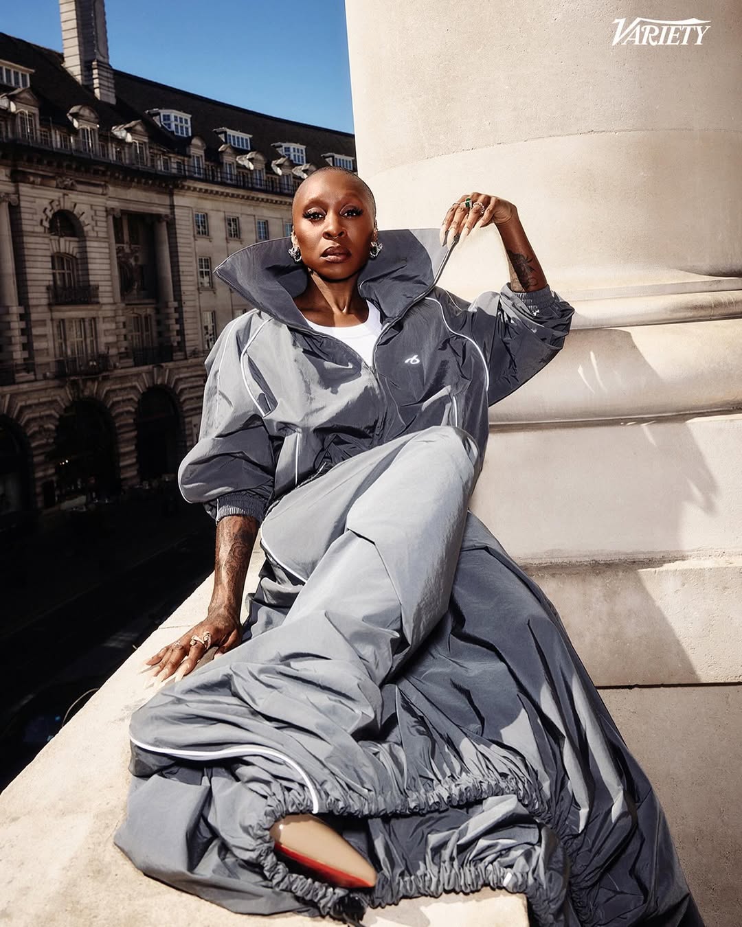 Cynthia Erivo wearing a grey windbreaker and long skirt seated on an architectural ledge outdoors with a classic European street background.