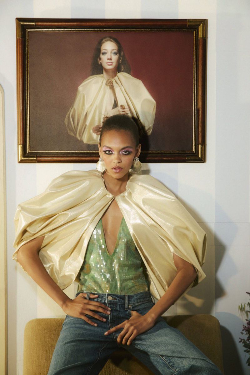 Model wearing a gold cape and green sequin top standing below a framed portrait painting by Carlijn Jacobs